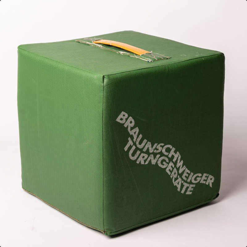 Ein grüner Sitzwürfel aus Turnmattenstoff mit der Schrift Braunschweiger Turngeräte und einem gelben Griff