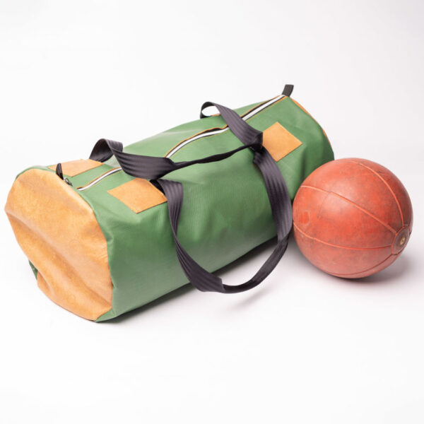 Eine grüne Tasche aus Turnmattenstoff und Tragegriffen aus Gurten von einem Auto. Daneben liegt ein brauner Medizinball.